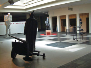 Exhibition view with large metal sculpture like a robot, grim reaper, a king and shopping person on a black and white carpet.